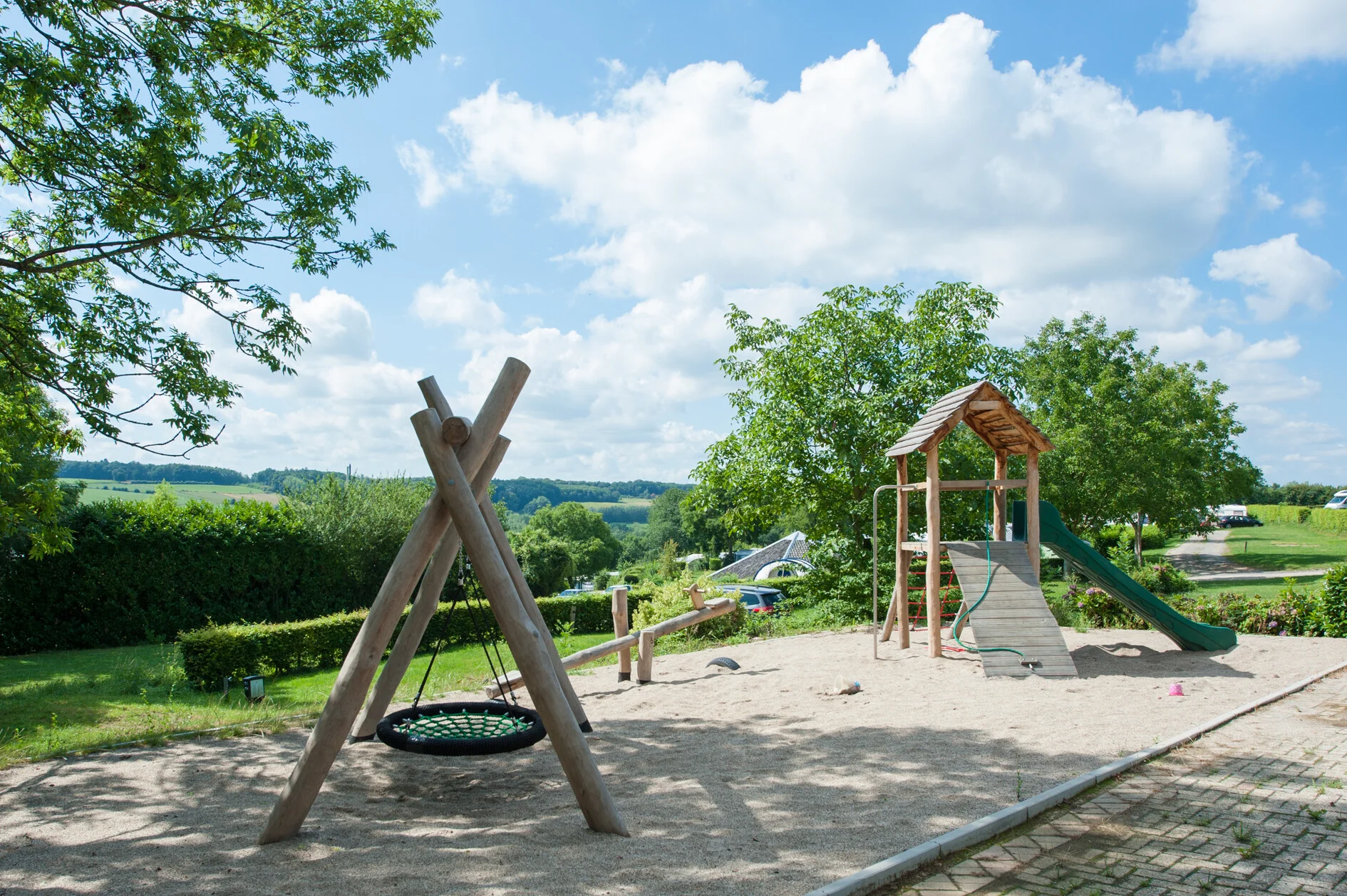 Speeltuin met nestschommel, wip en houten speeltoestel op het campingterrein met heuvels op de achtergrond